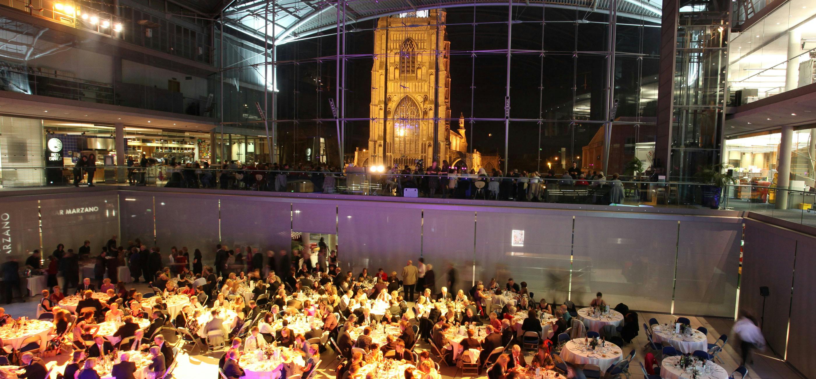 A formal event with tables in The Atrium of The Forum in the evening with St Peter Mancroft lit up outside.