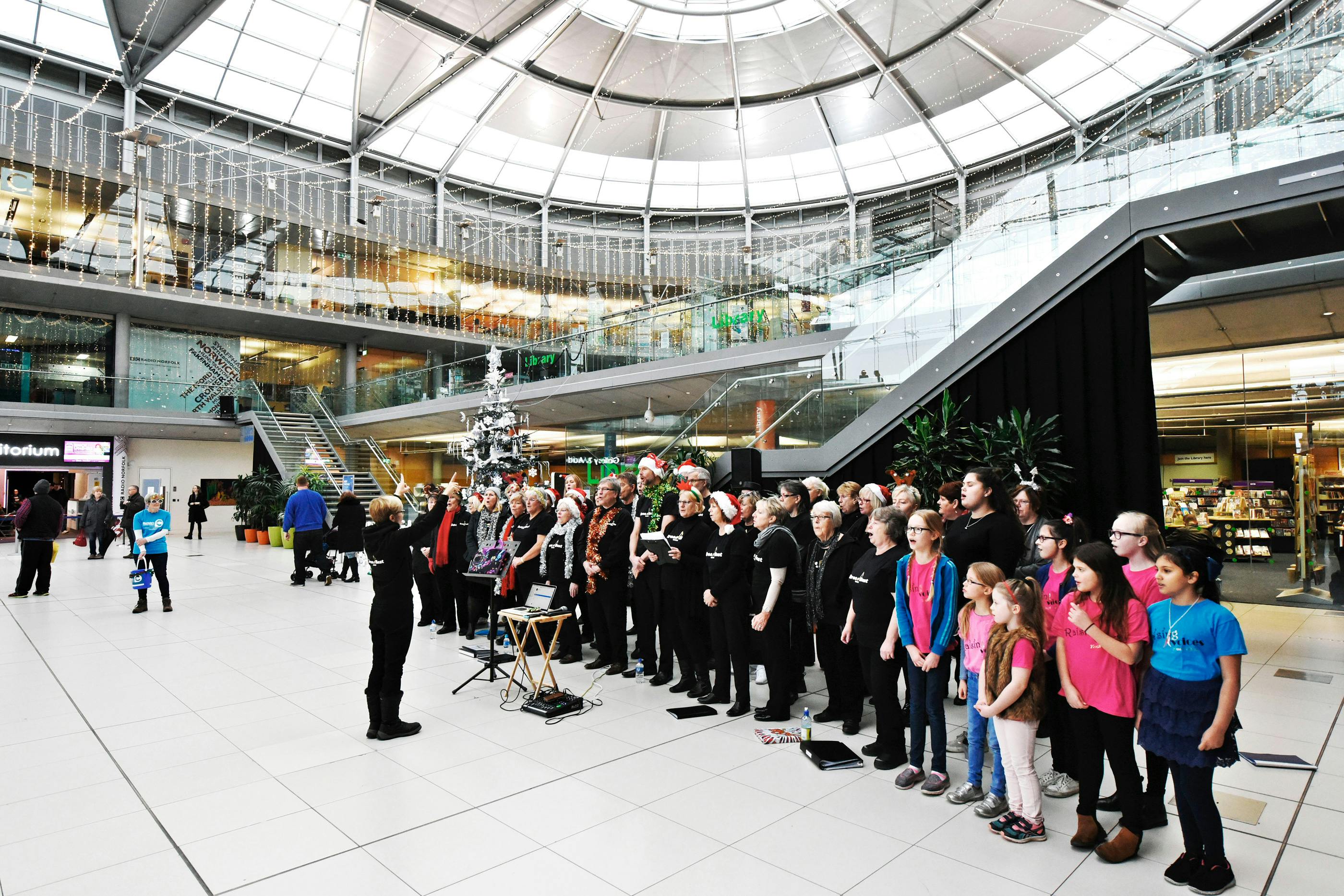A conductor and choir sing in the Atrium of The Forum dressed in festive hats and tinsel