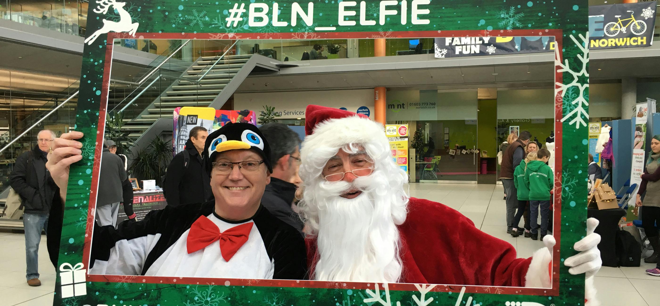 Two people take a selfie in a festive photo frame, one is dressed as a penguin, one as Father Christmas