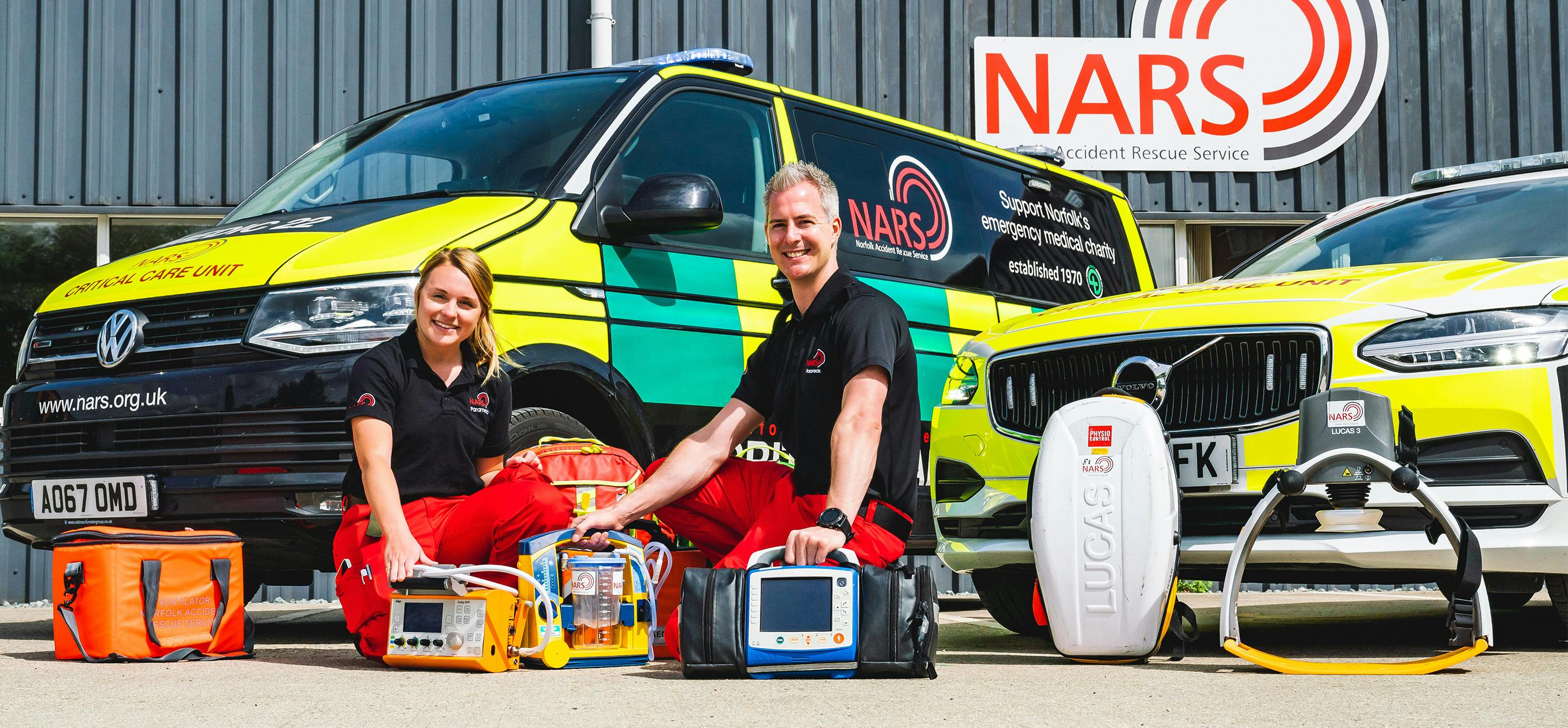 Two members of the Norfolk Lowland Search and Rescue crouch next to their emergency vehicles with various items of emergency equipment in front of them.