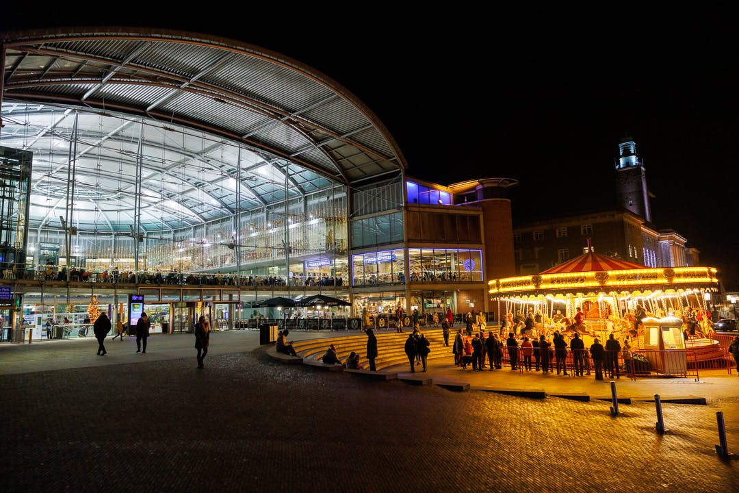 The image shows The Forum building exterior at night, the interior adorned with Christmas Fairy lights, outside shows silhouettes of people around the traditional classic carousel, with Norwich city hall in the background