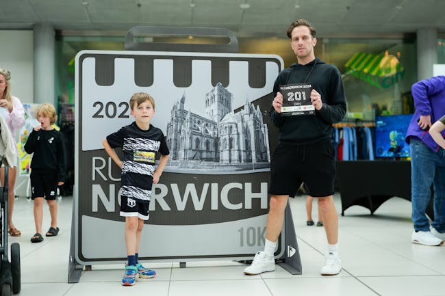 A man and a boy dressed in running gear stand triumphantly in front of a Run Norwich sign.