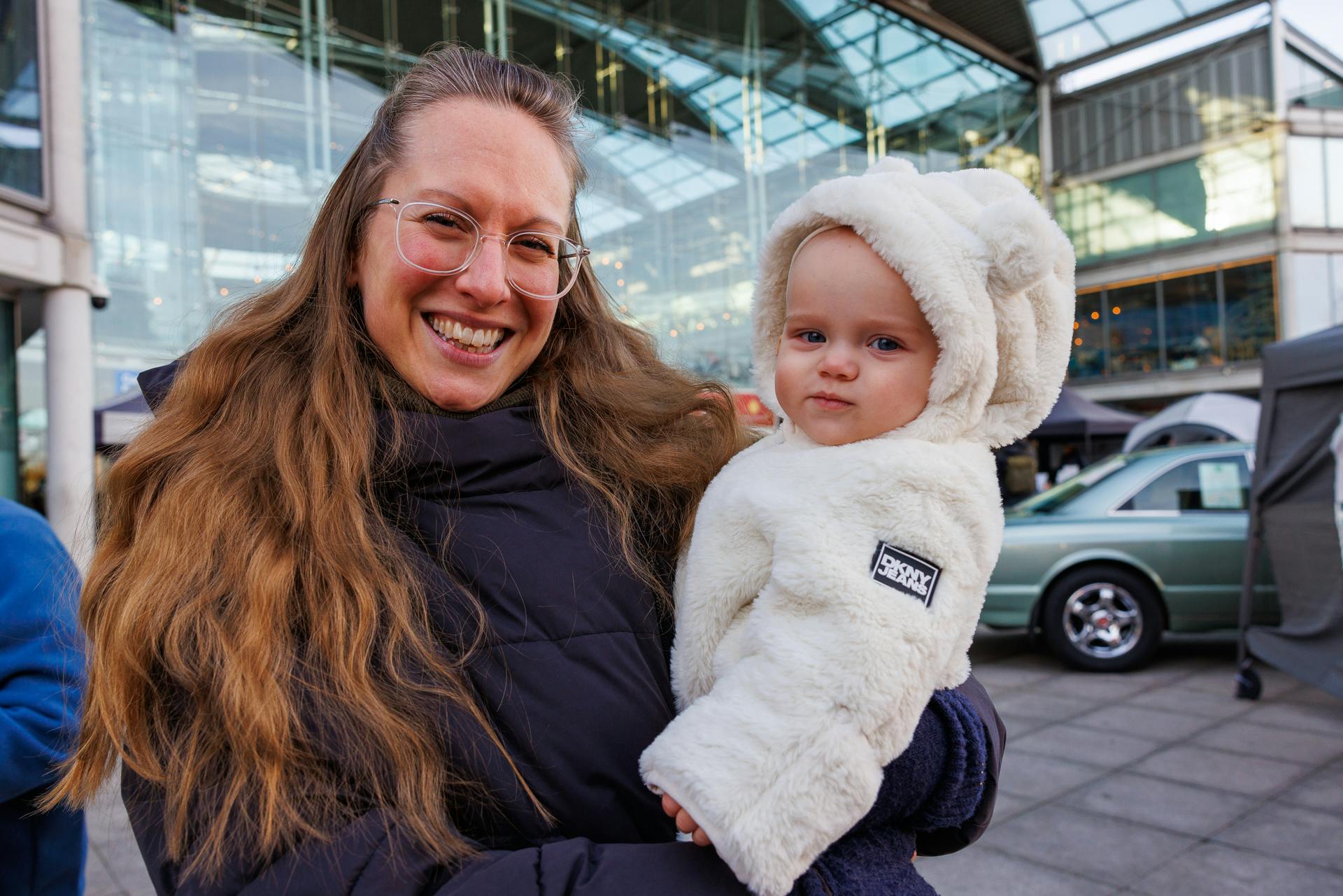A woman with glasses and long brown/blonde hair grins at the camera and is holding a small child with a white fluffy suit and hood.