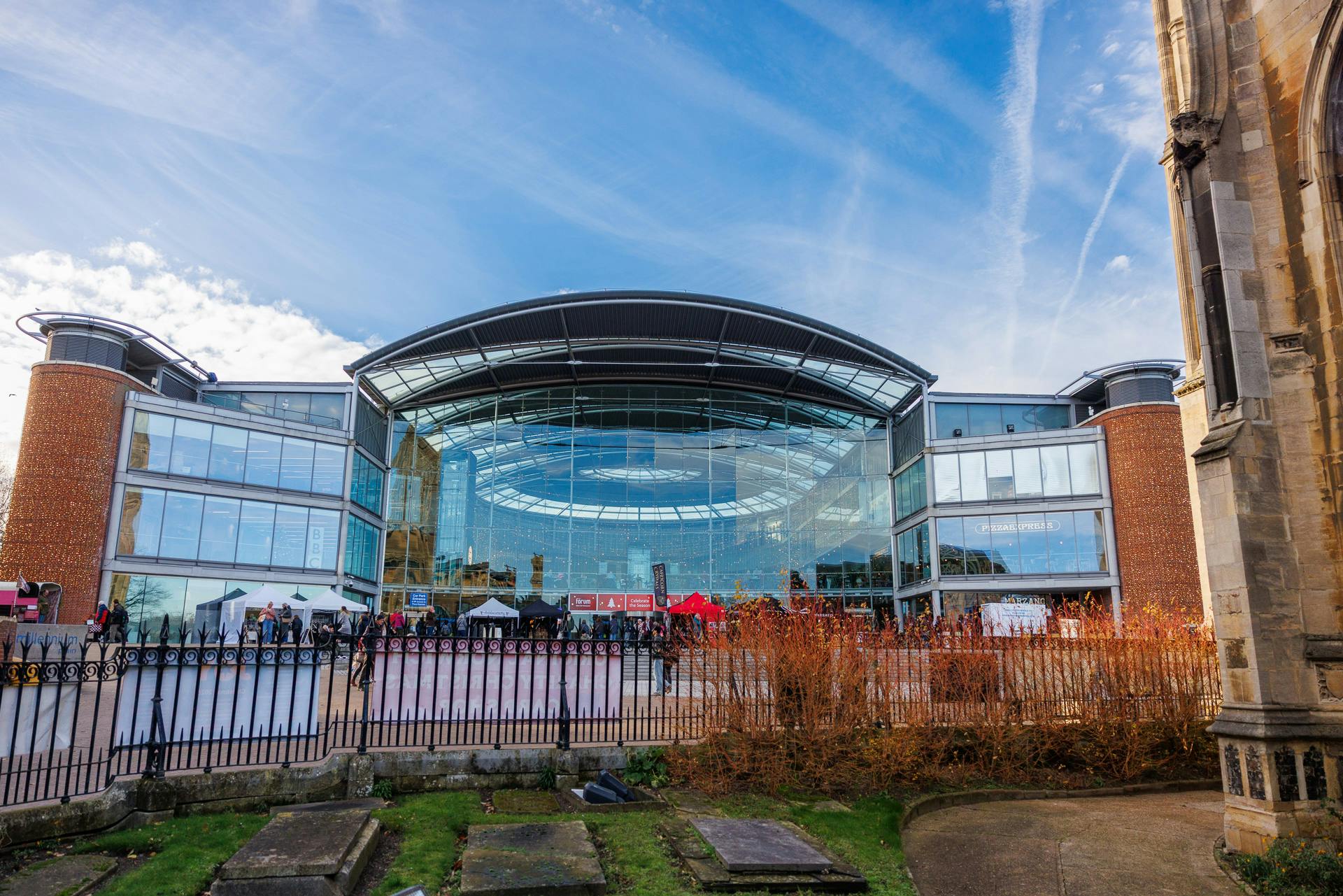 forum_5_12_25_photos-117.jpg View of the front exterior of The Forum, from the garden of St Peter Mancroft church, in front of a blue sky with wispy clouds.
