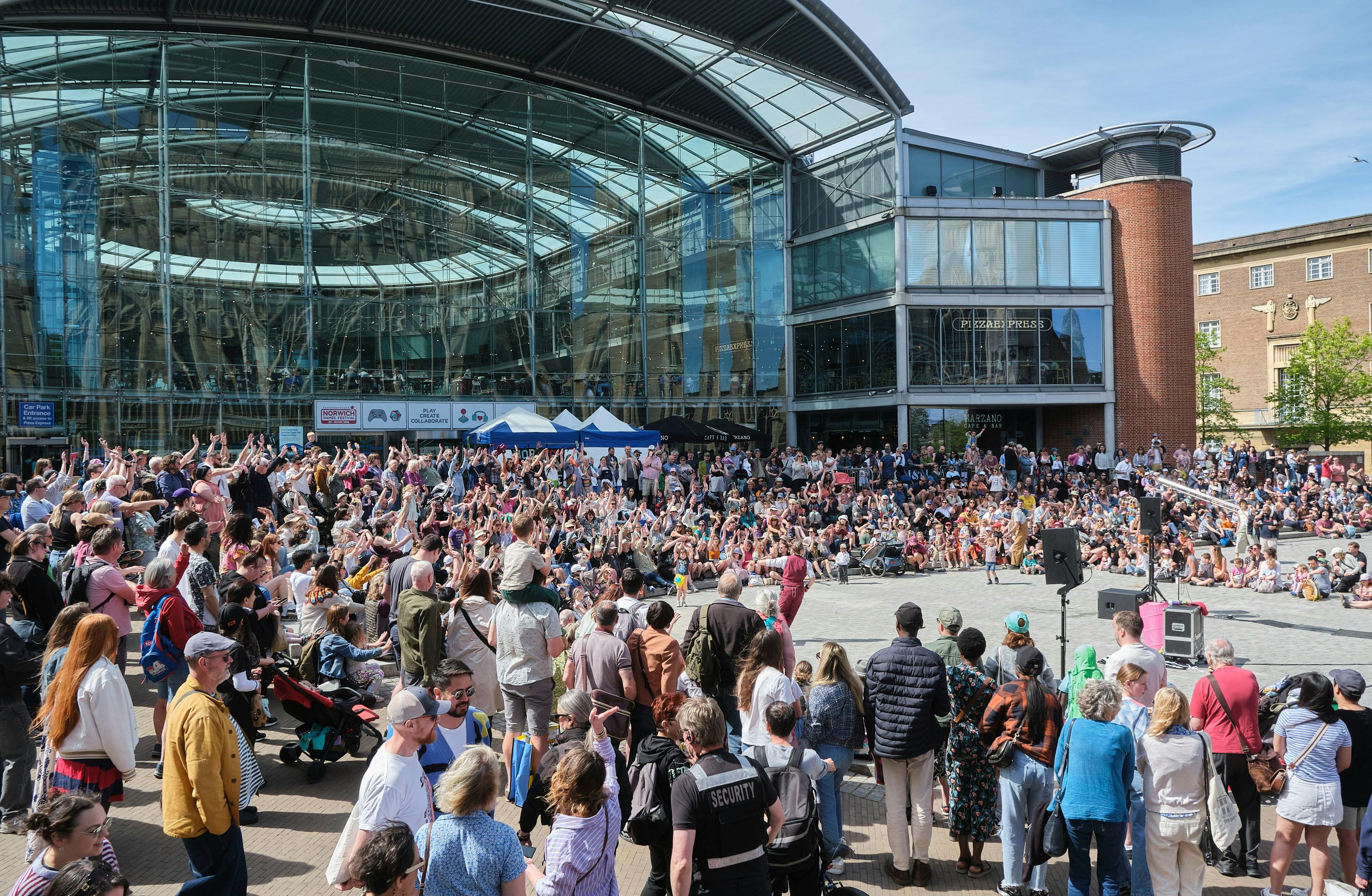 The exterior of The Forum building, with a crowd of people watching performance in the Amphitheatre. the crowd is doing a mexican wave, it is a bright, sunny day.