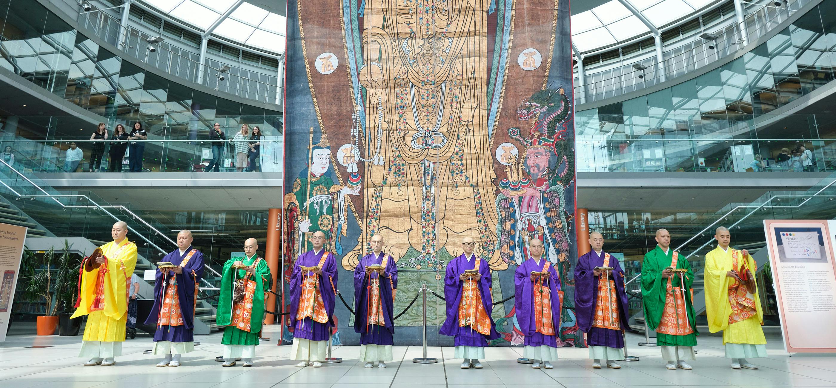 A row on monks in colourful robes stand in front of a large banner with Buddhist imagery