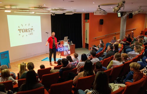 A live poetry performance in a modern auditorium space with tiered seating.