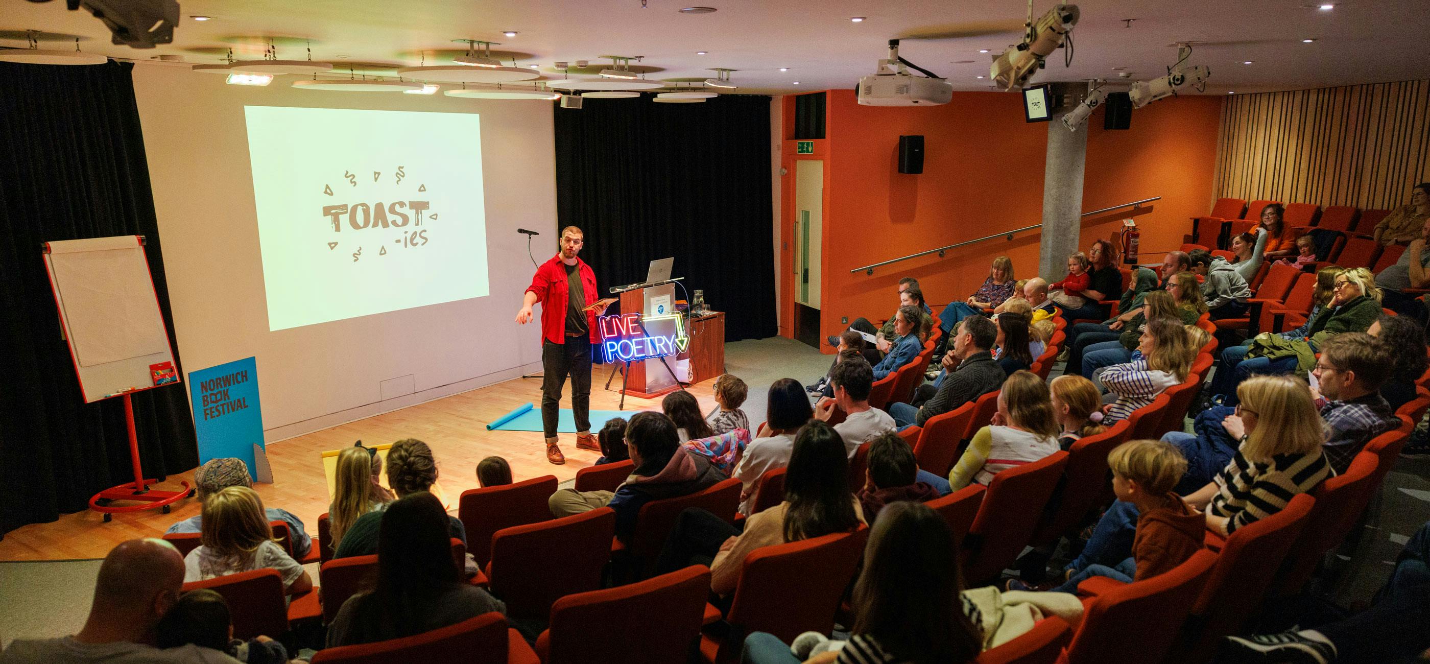A live poetry performance in a modern auditorium space with tiered seating.