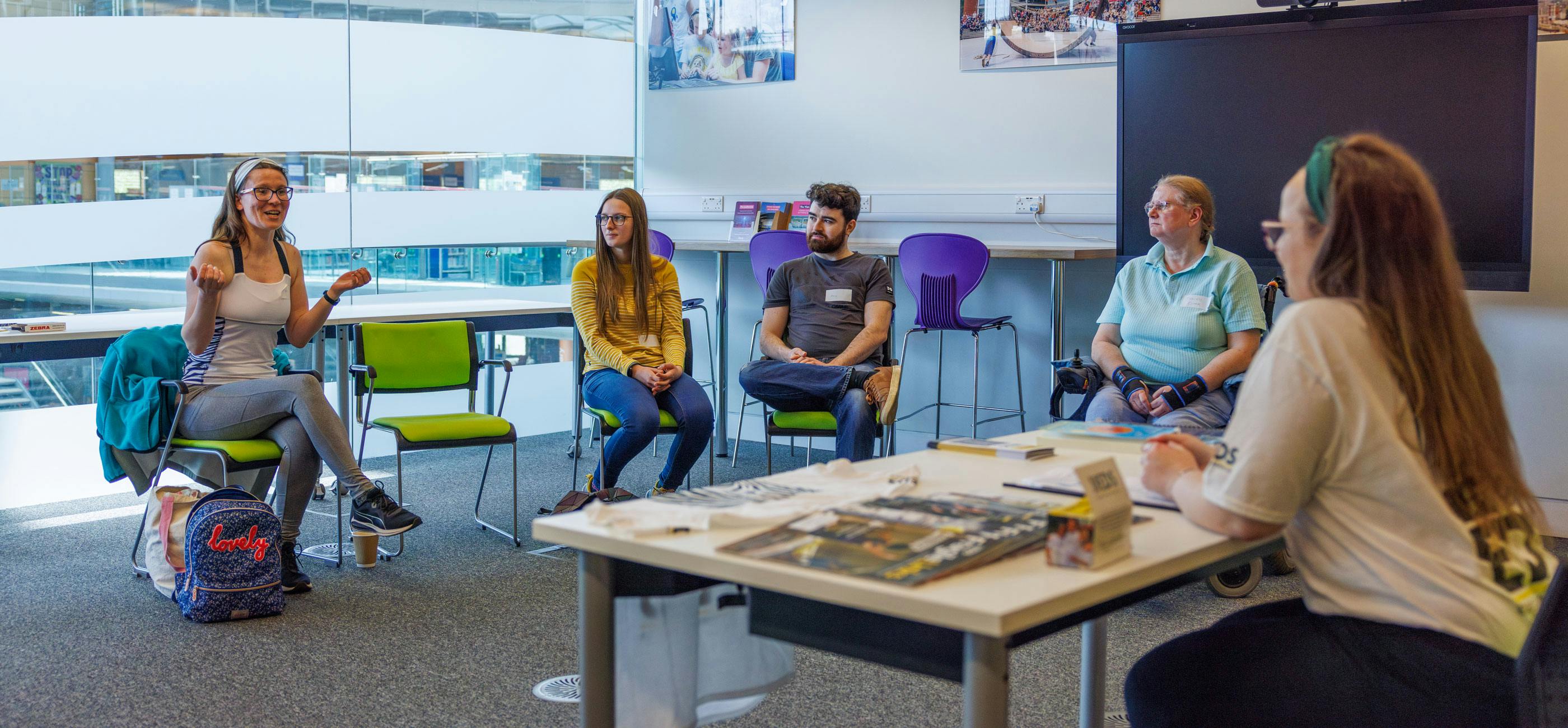 A group of people sit in a light filled meeting room having a discussion.