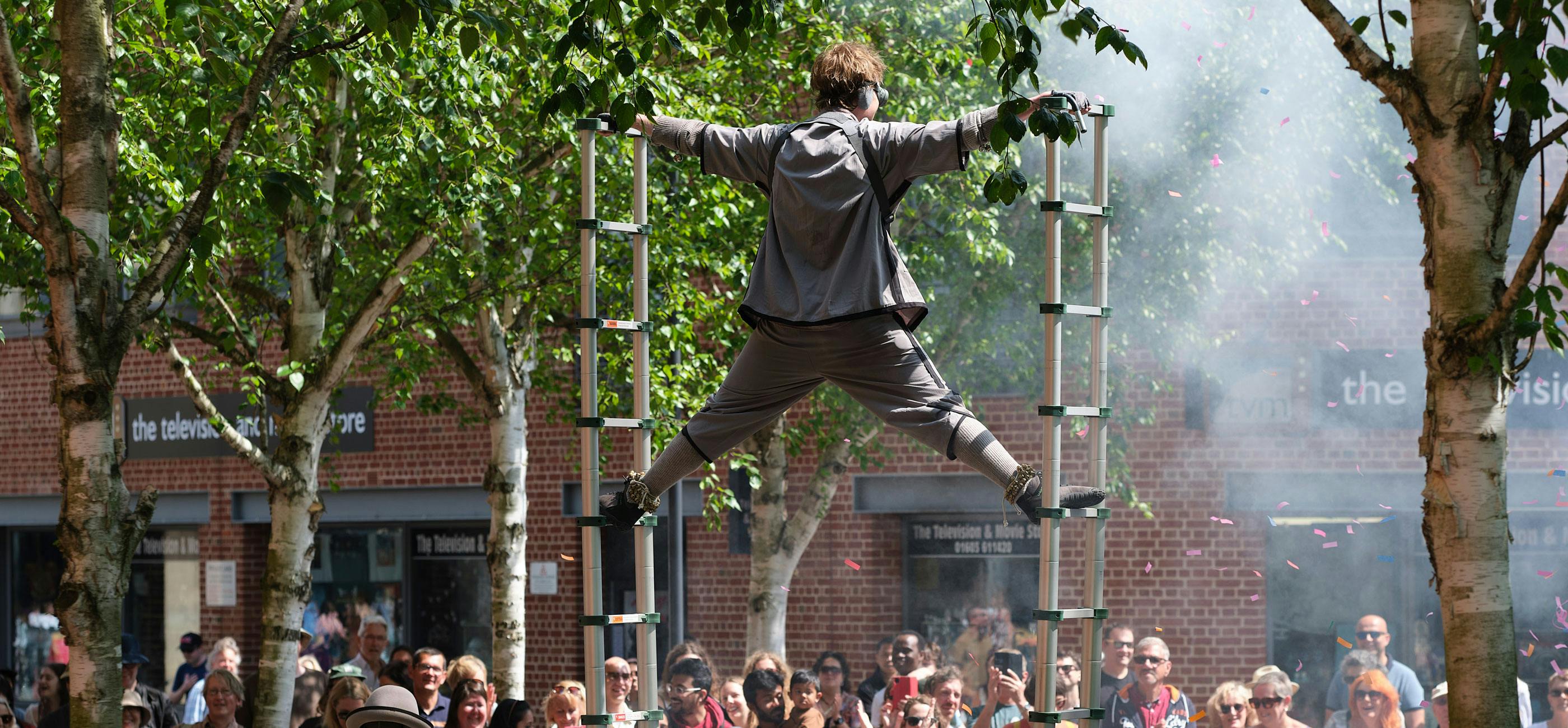 A performer on stilts, with a crowd watching and smiling on Theatre Plain