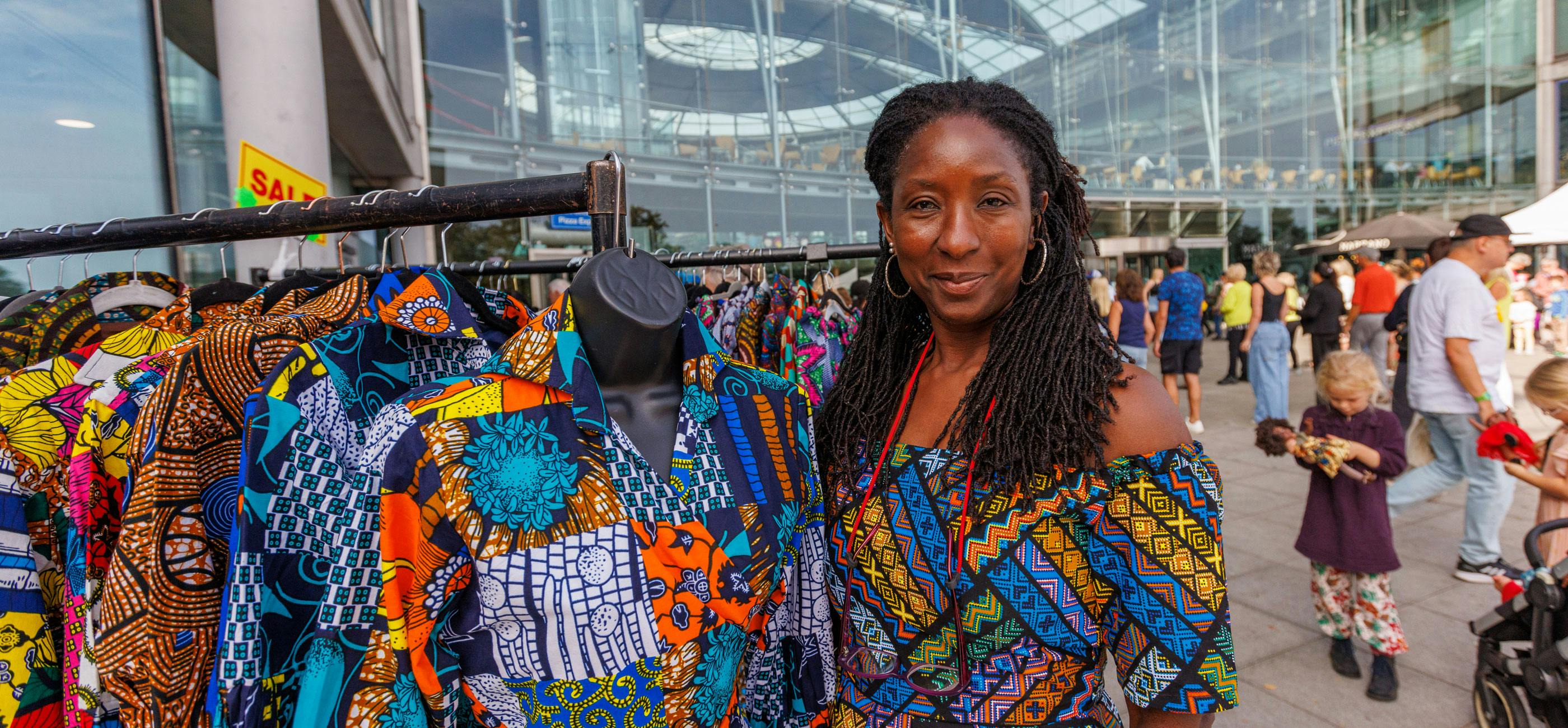 A stall holder smiles outside The Forum in Norwich, with a rail of colourful, African style print clothes..
