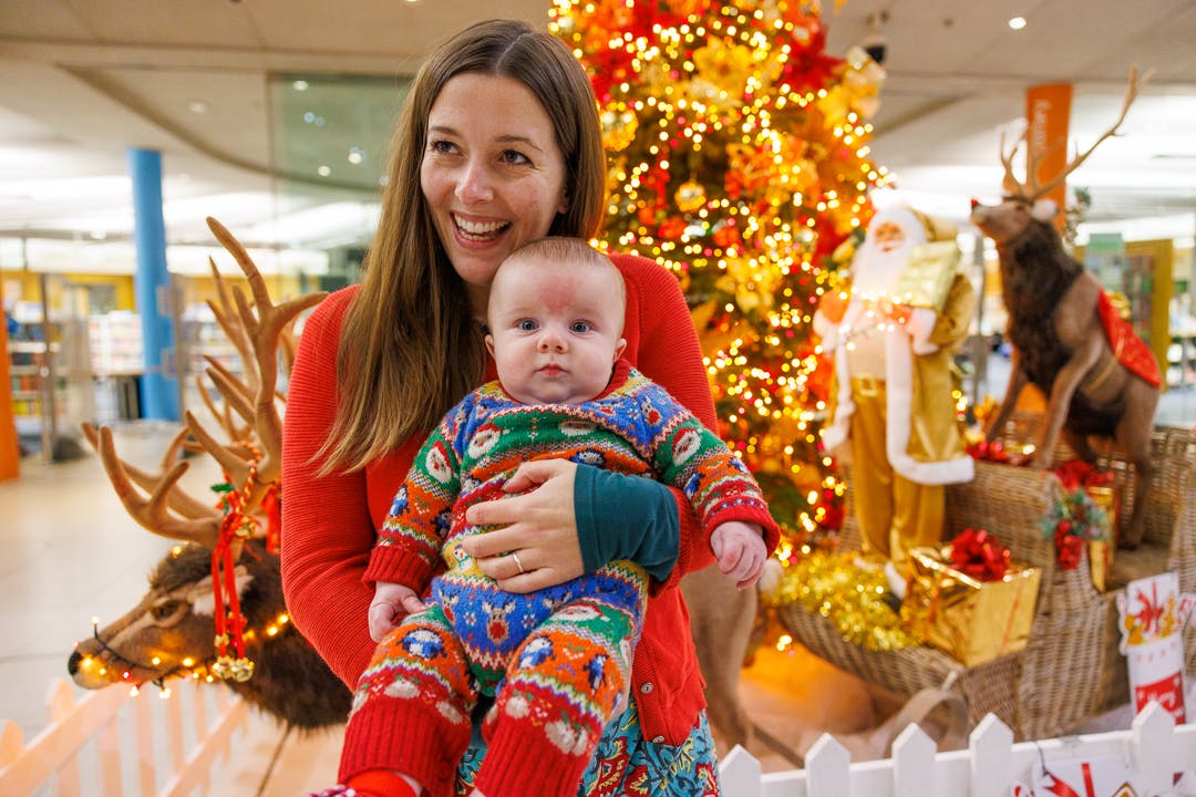 A woman stands in front of The Forum Christmas Tree decorated with red and gold ornaments and sparkly golden lights. She is holding a baby in a knitted christmas onesie.