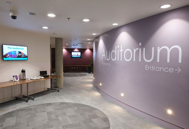 A modern lobby area with tables, digital screens and a large sign showing the entrance to the auditorium