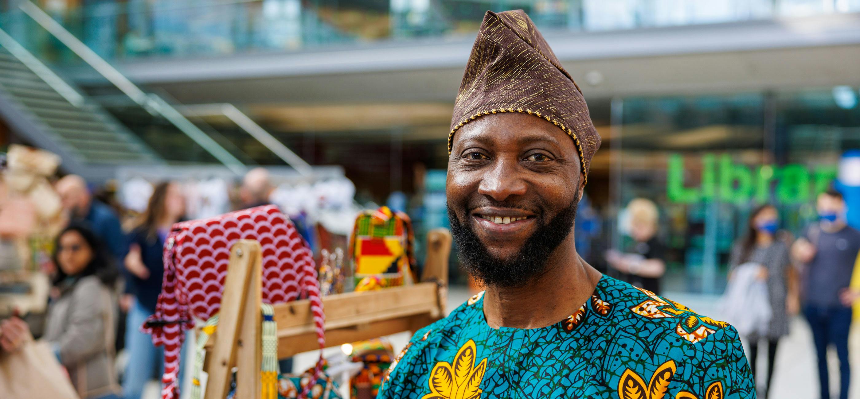 A smiling market stand trader stands inside The Forum dressed and selling accessories in vibrant African fabrics.