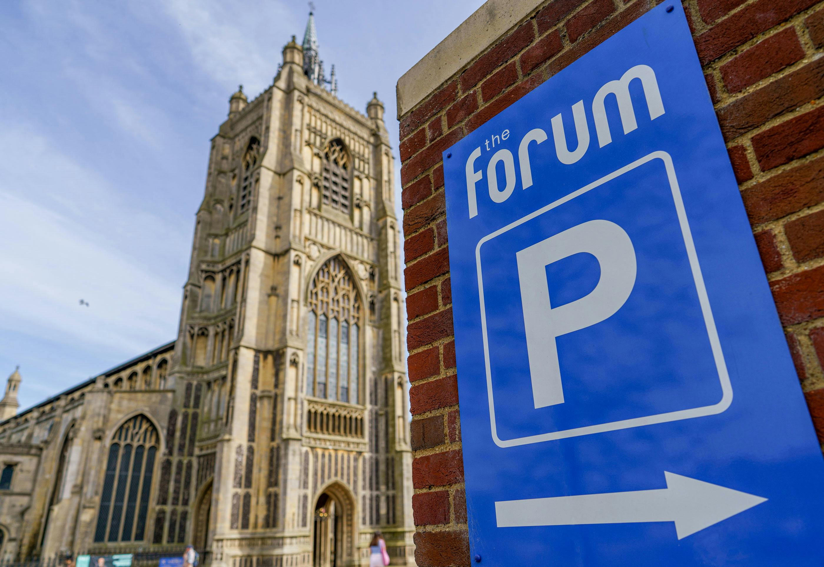 A blue sign with The Forum, a parking symbol and an arrow pointing to the right with St Peter Mancroft church in the distance.