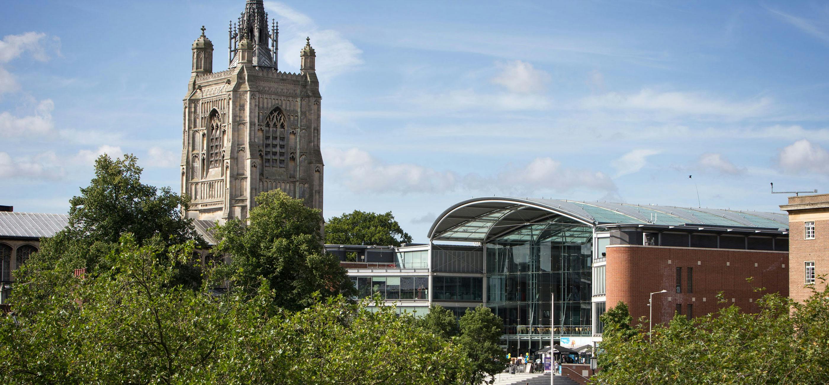 A wide shot over Norwich Market of The Forum, a modern glass and red brick building and the spire of St Peter Mancroft Church.