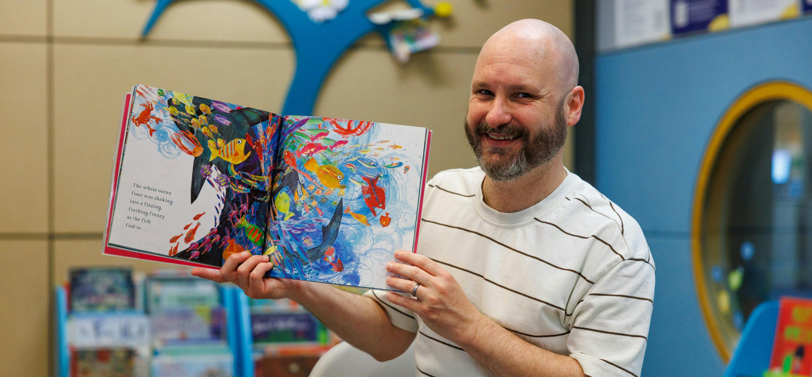Librarian Adam smiling holding up a colourful children's book in the Millennium Library