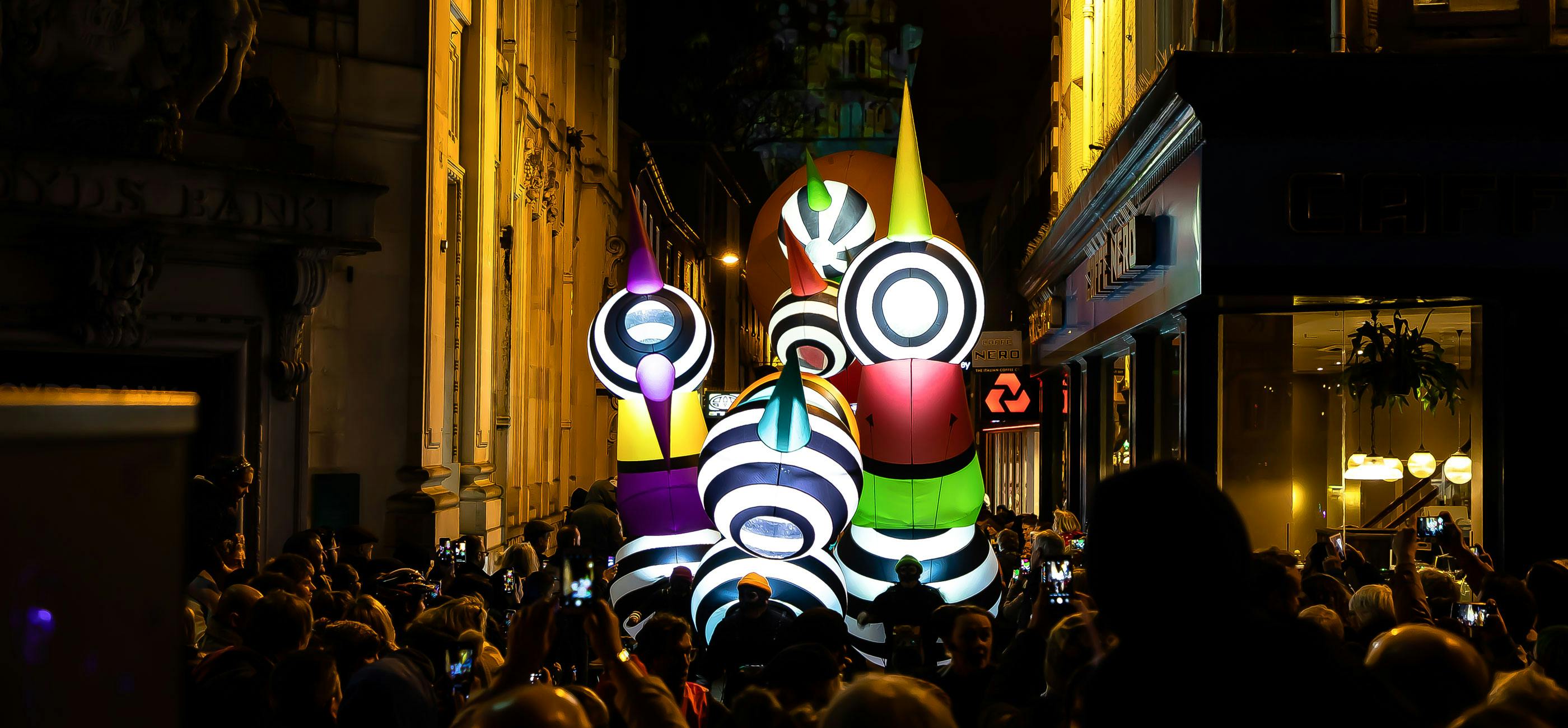 A colourful light-up inflatable parades through the streets of Norwich at night surrounded by a crowd.