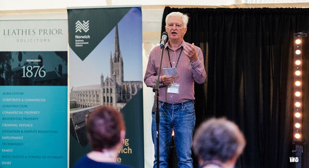 A man speaks on a stage with Norwich BID banner in the background.