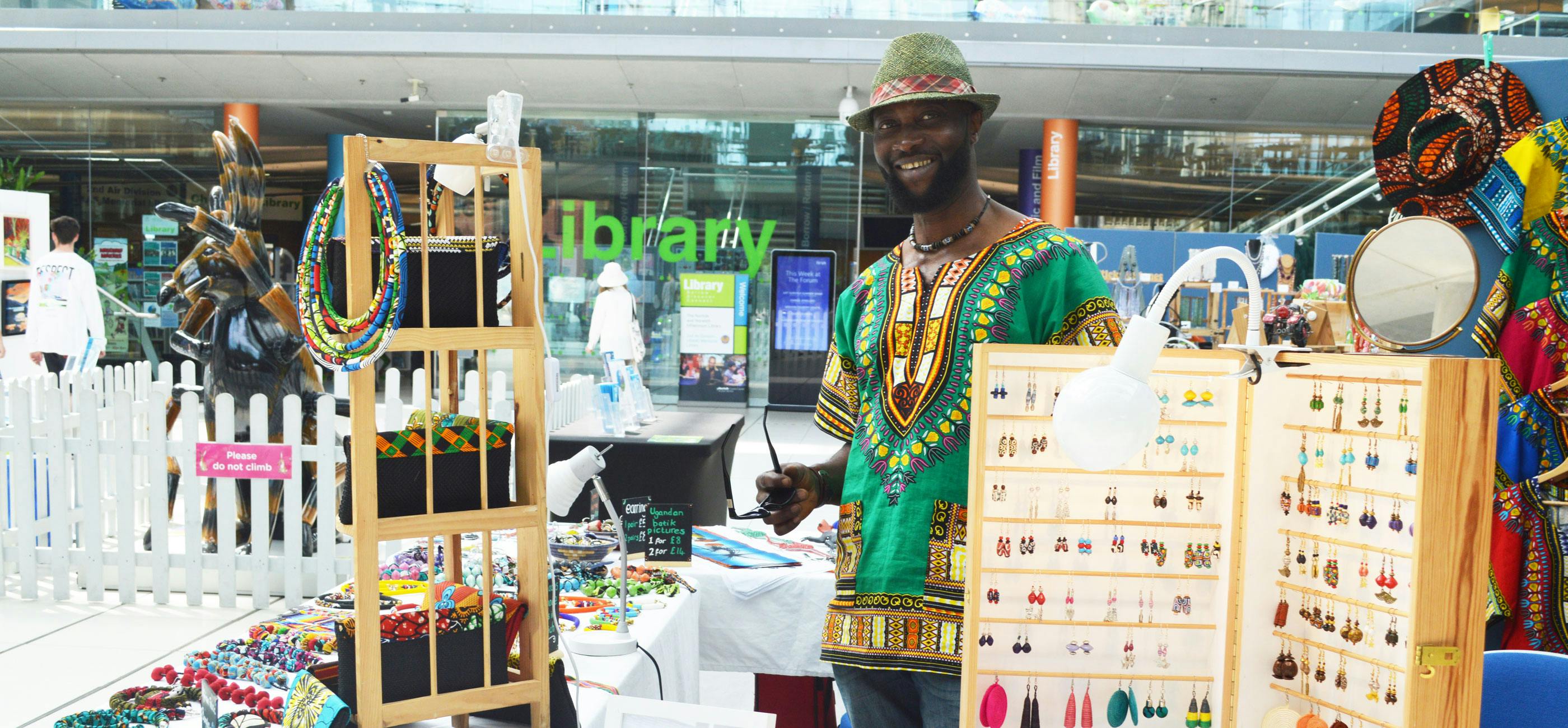 A trader at a craft market stands smiling at his stand which has colourful, beaded jewellery for sale.