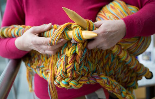 A close up of someone knitting with large knitting needles with yellow, orange, red and turquoise wool.