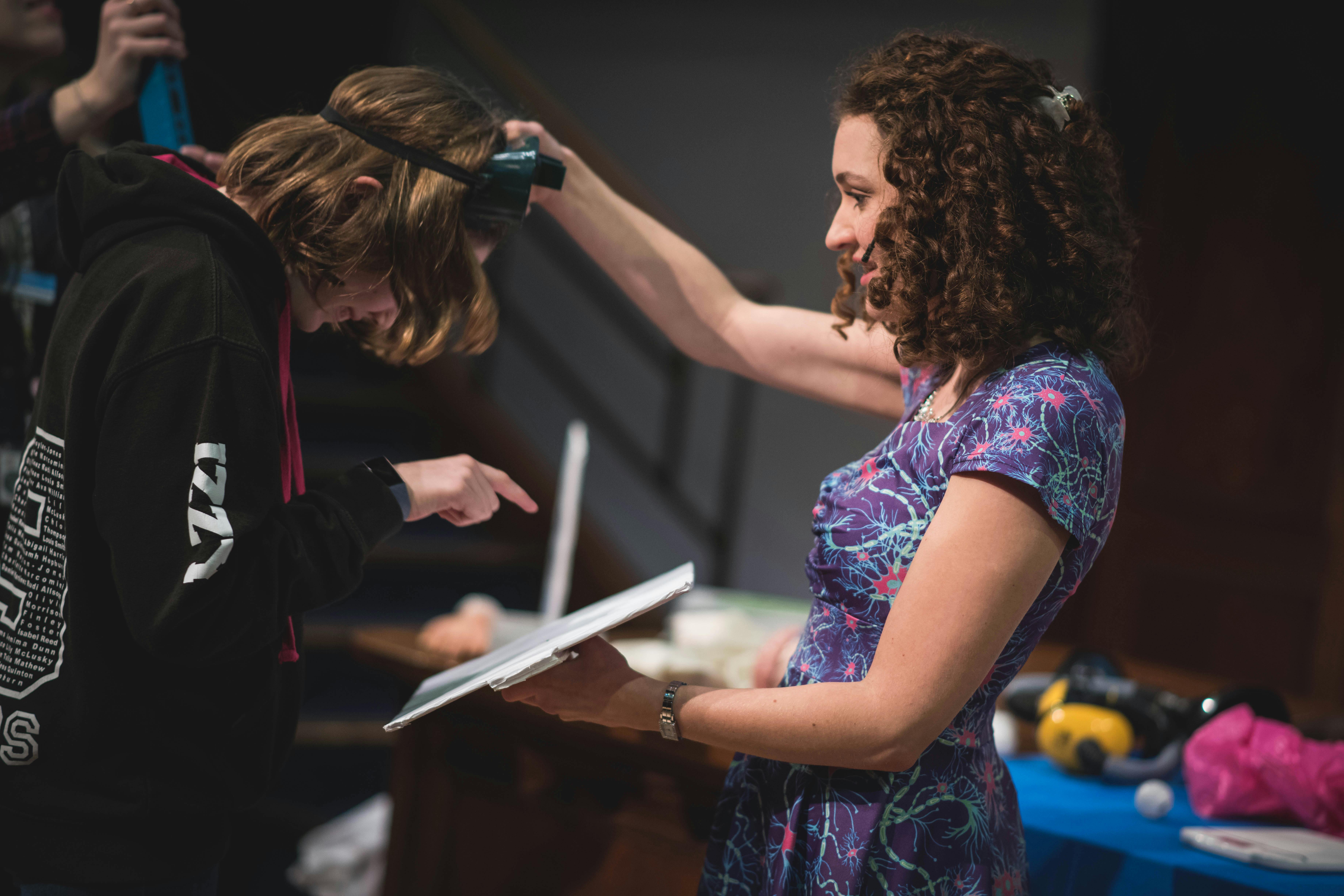 braintastic-goggles.jpg Woman in science-themed dress holding goggles up to a student as they read