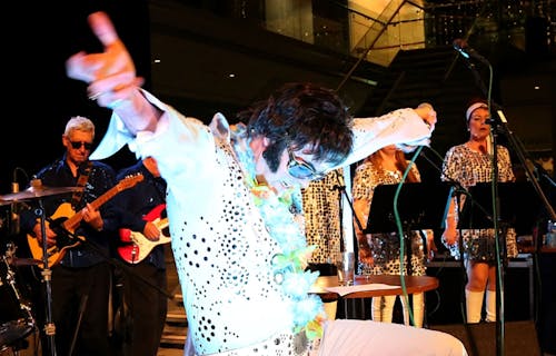 An Elvis impersonator, in white shirt and glasses, performs in front of a band.