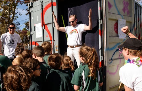 A group of school children stand in front of smiling performers with colourful 10 minute dance party t-shirts on. They're standing in front of a shipping container decorated in colourful graffiti.