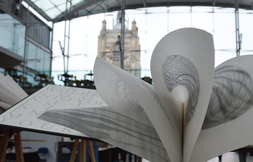 A book folded open with fine pencil drawings, the glass front of The Forum is in the background.