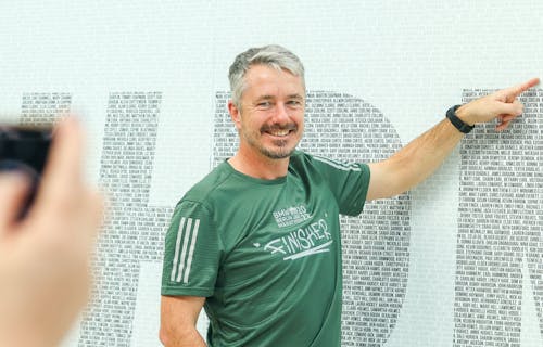 A runner points at his name on a wall with thousands of participants names printed.