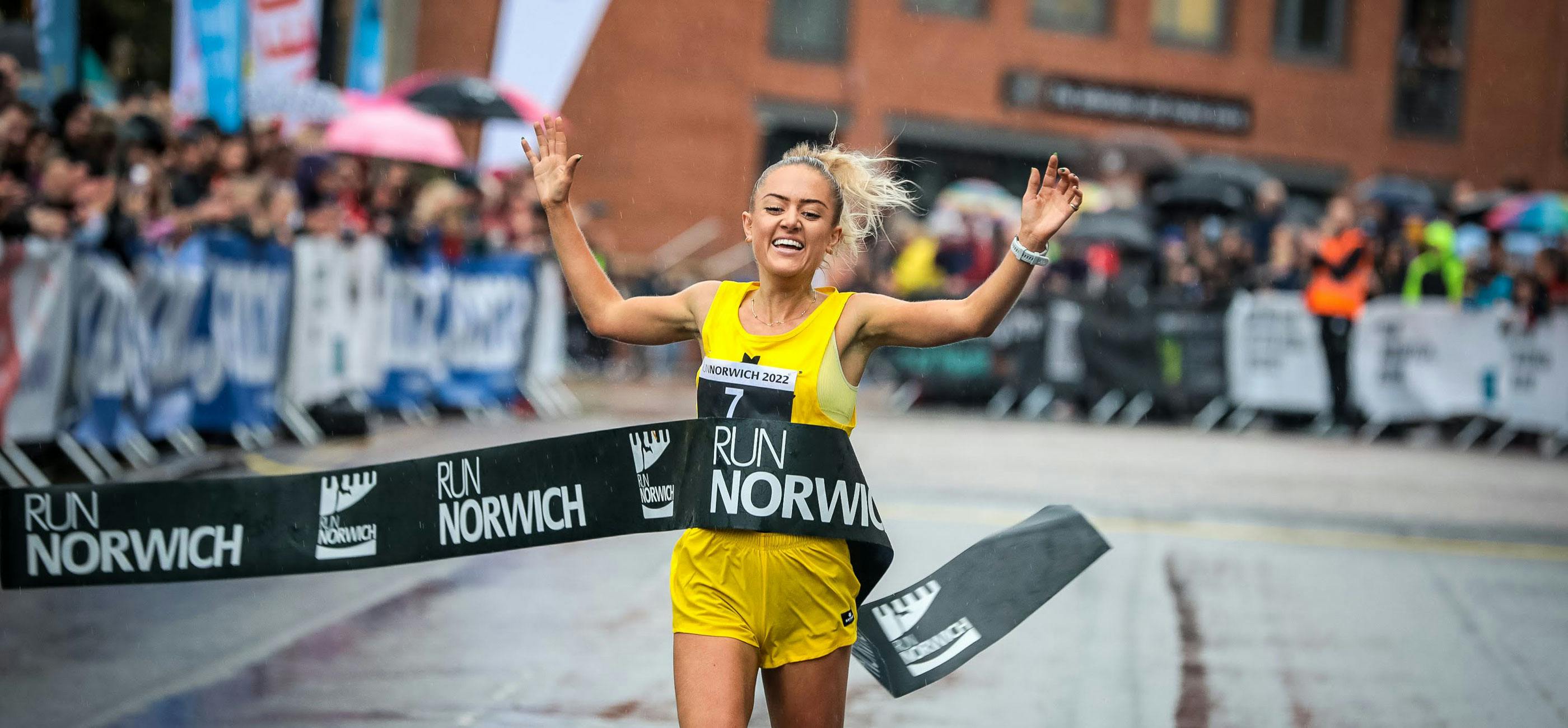 A woman in a yellow runs through the Run Norwich finish line ribbon smiling.