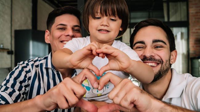 Two dads and their child make heart shapes with their hands
