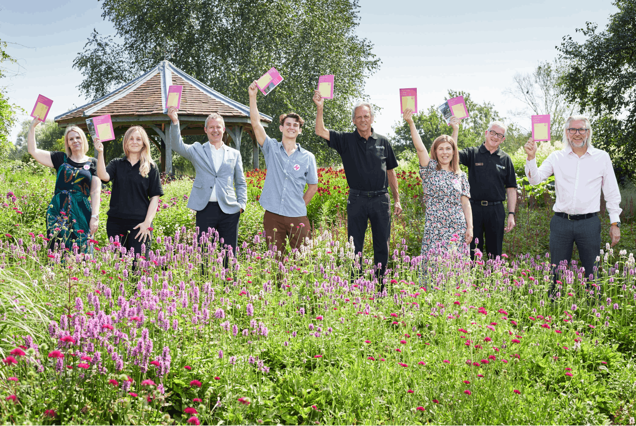 Members of the Norfolk Heritage Open Days and Pensthorpe team hold up the 2021 brochure in a park full of flowers.