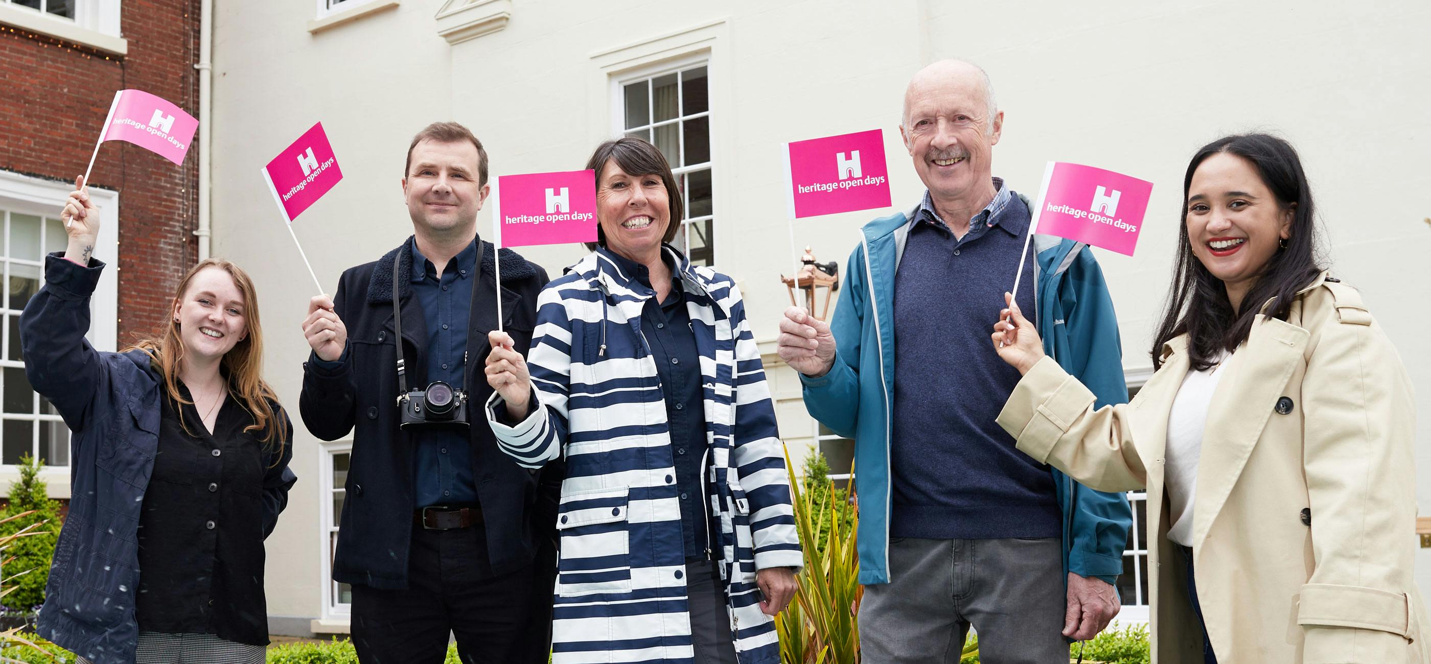 A group of people stand waving pink Heritage Open Days flags smiling.