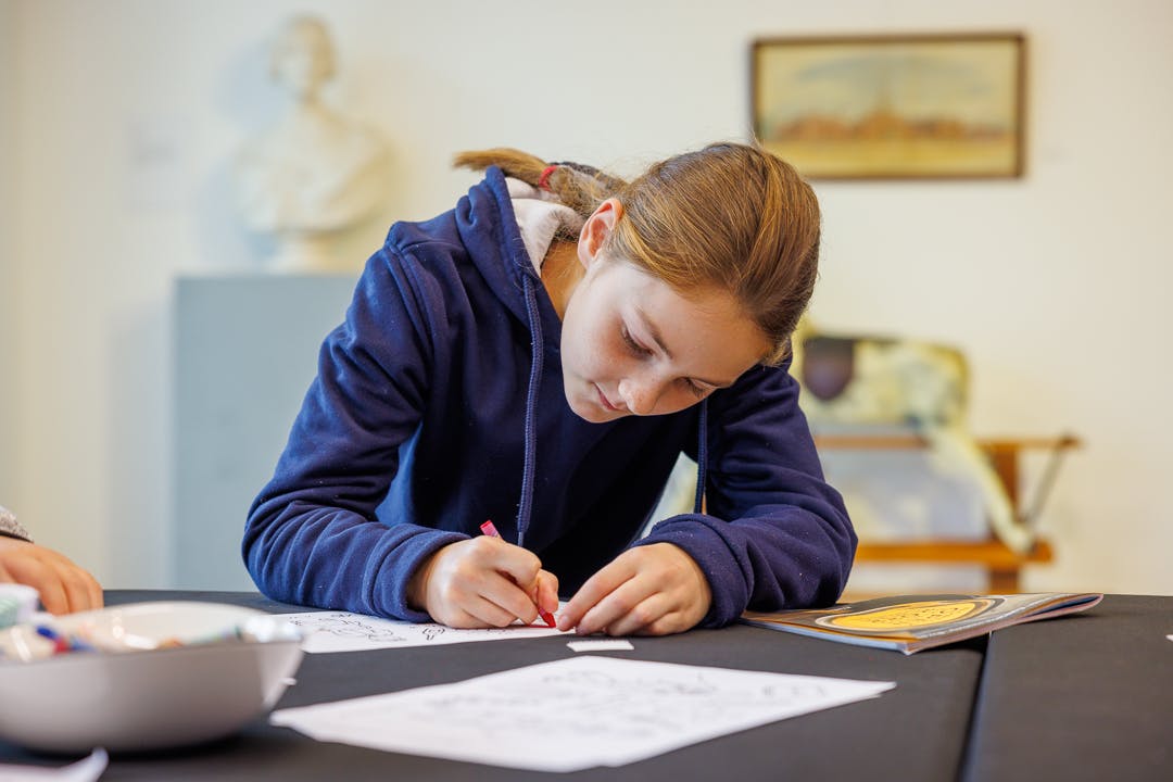 A young girl in dark blue hoodie leans over a table to colour in a picture in front of a statue, painting and rocking horse
