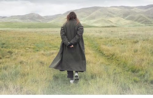 photo of a woman in a long coat walking toward some hills