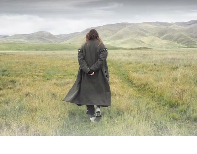 photo of a woman in a long coat walking toward some hills