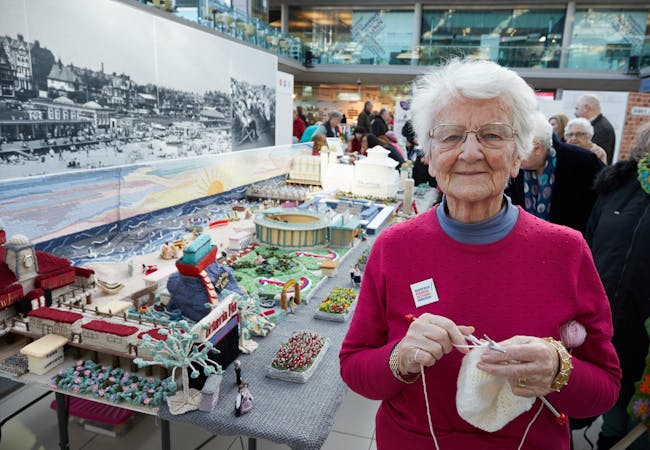 Elderly lady smiling in front of a knitted display