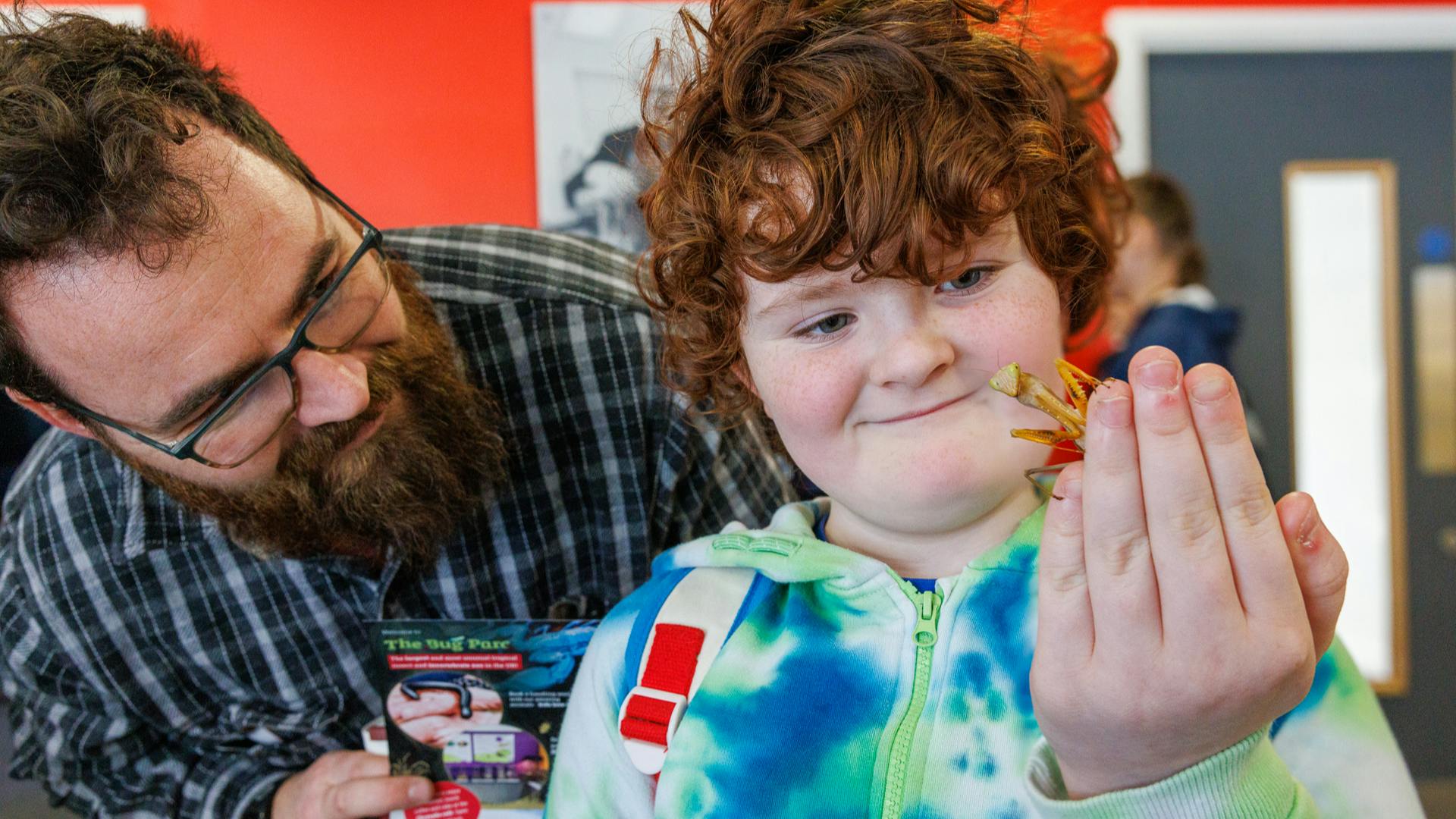 A child holds a grasshopper in their hand whilst an adult looks on