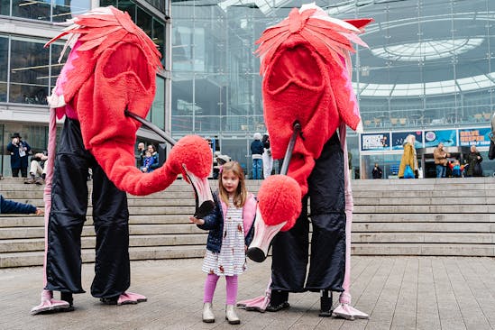 A child stands next to two large flamingo puppets outside The Forum
