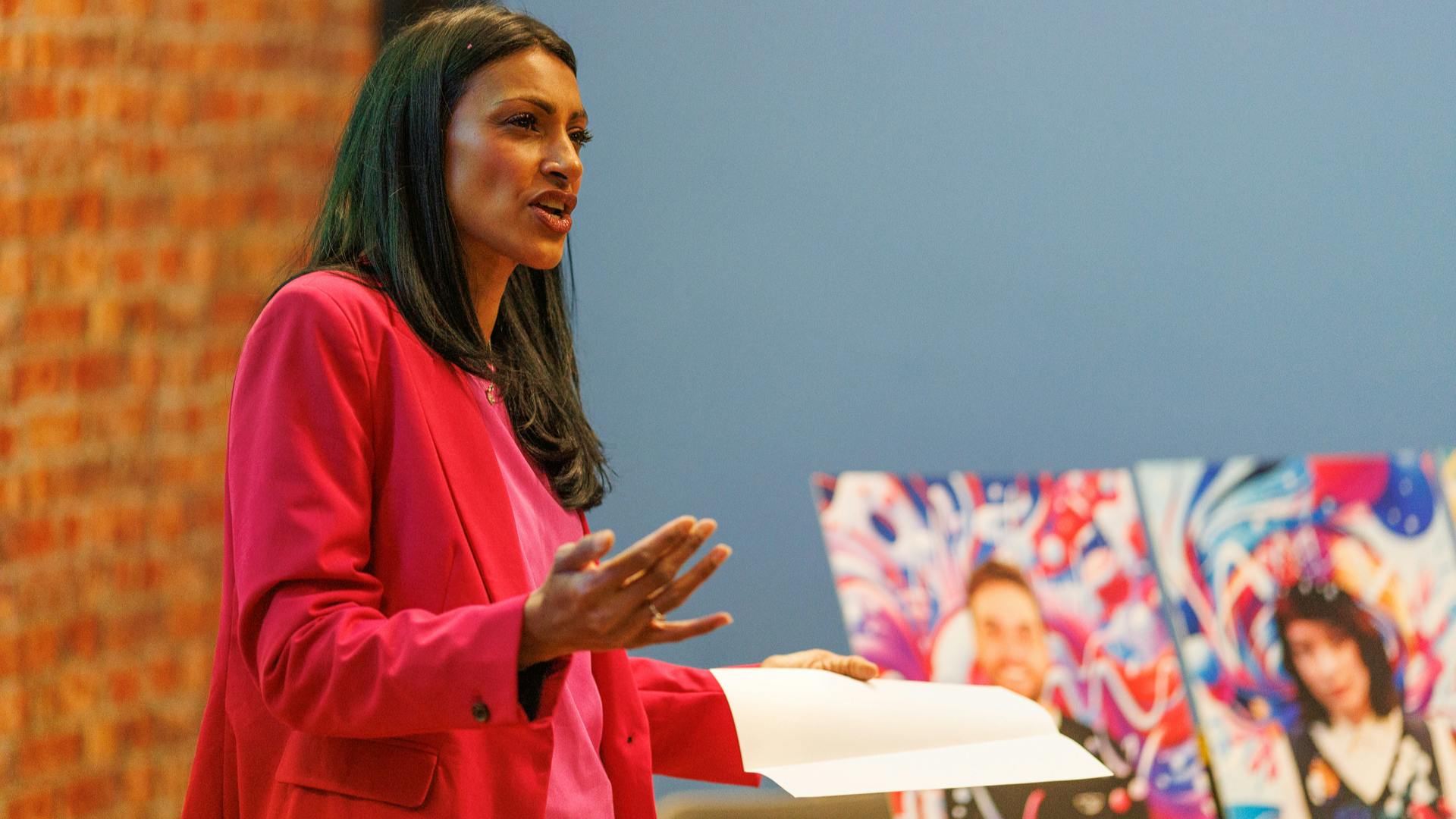 A woman with long dark hair and wearing a pink suit speaks to an audience.
