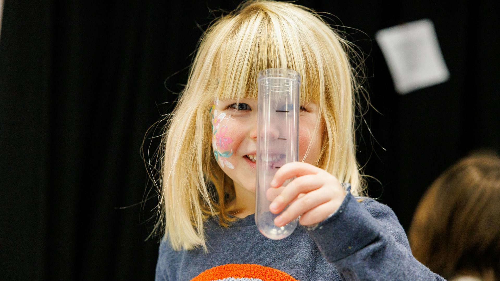 Little girl with face paint on her cheek holds up a plastic test tube