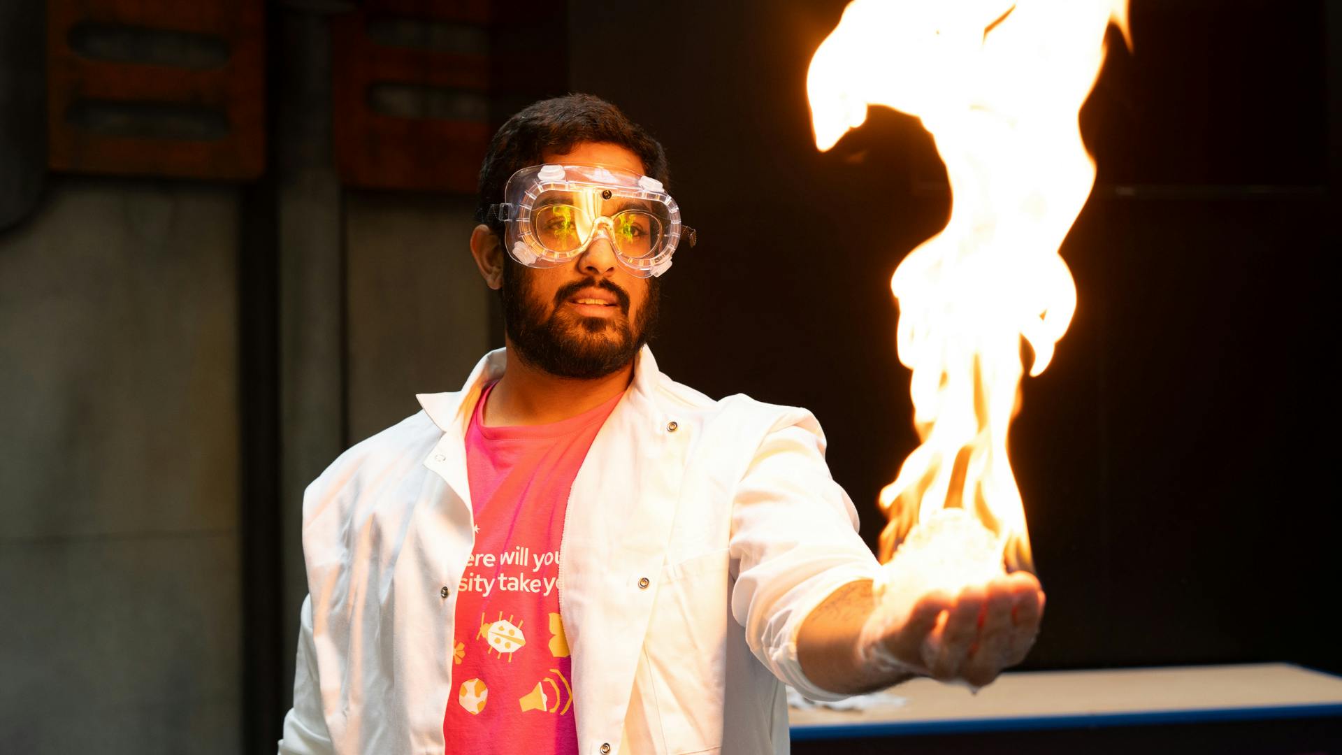 A person wearing safety goggles and a lab coat holds out their hand which is lit with a bright flame.