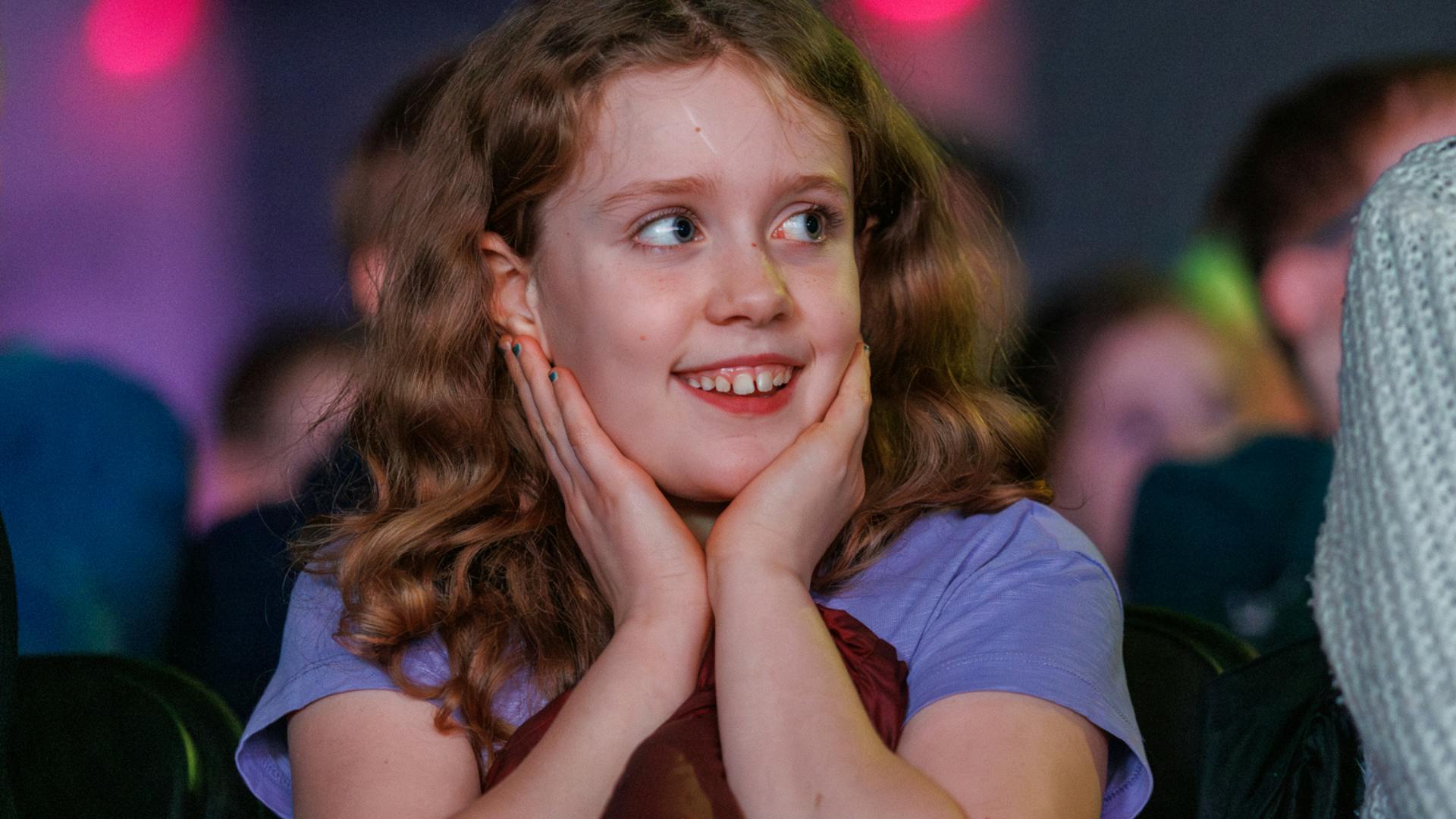 A child in an audience smiles and looks on in wonder, with hands on their face.