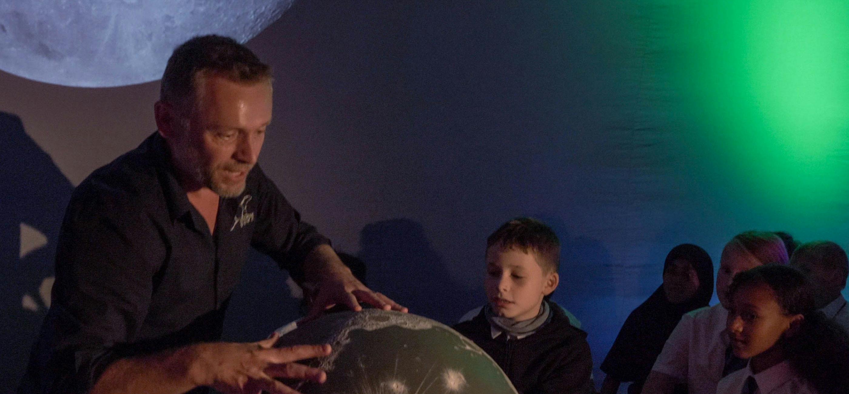Inside the Explorer Dome, with a man with hands on a model moon, with the moon projected onto the inside of the dome, with children watching