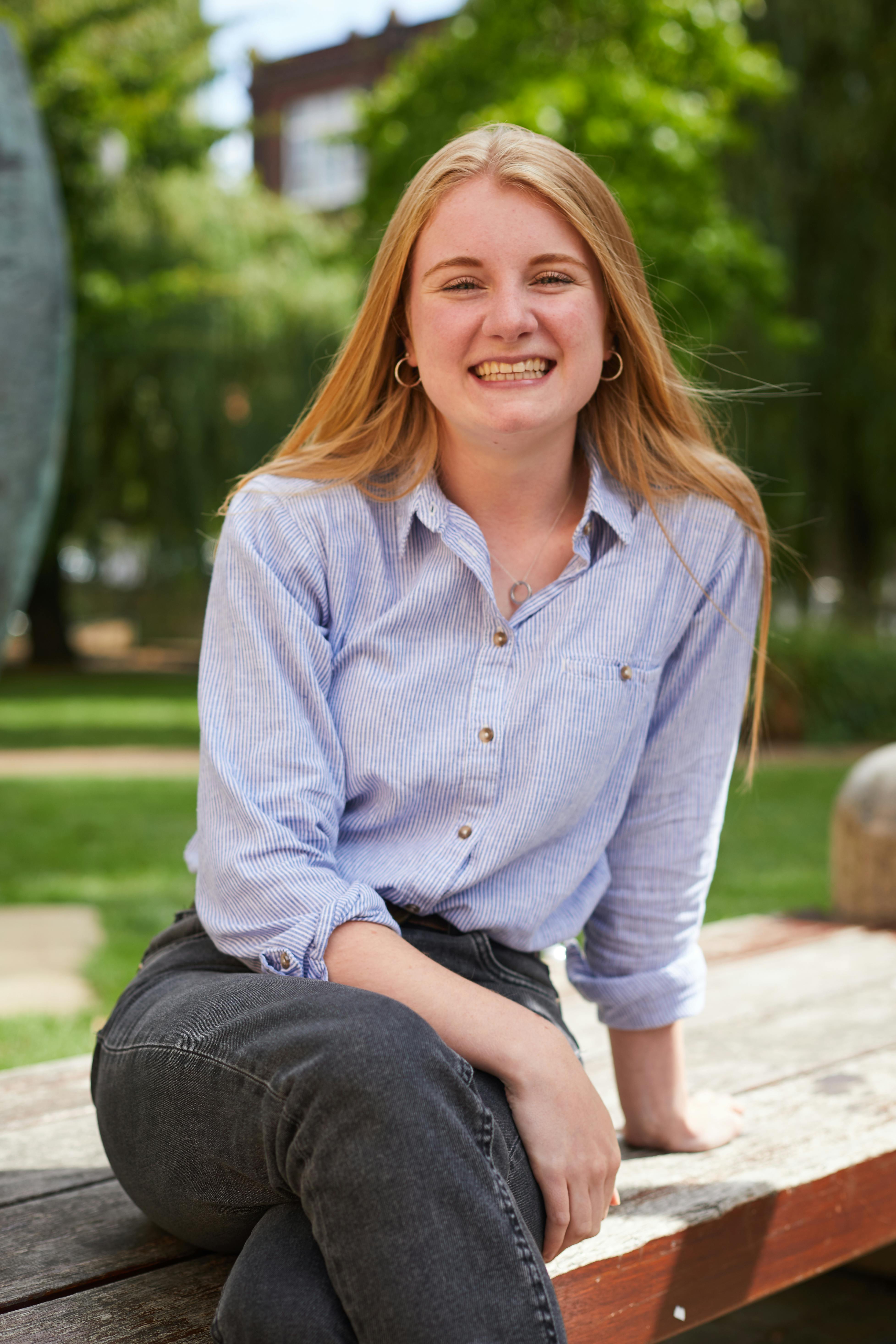 Hannah Linsdell sat on a bench in a park, looking at camera smiling, wearing striped blue shirt, black jeans, hoop earrings