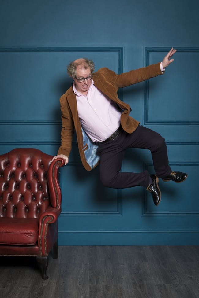 Image of Peter Lovatt dancing - doing a heel kick whilst leaning on a red leather chair, with blue wall in background