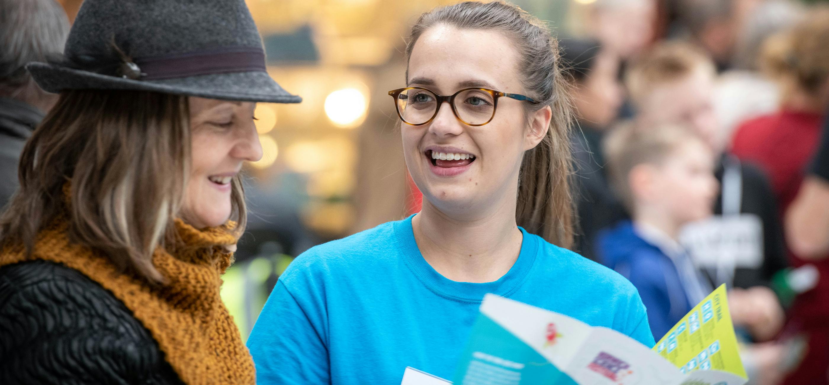 NSF volunteer in a blue tee shirt and glasses, shows a trail map to a member of the public, in a black coat, yellow scarf and grey hat