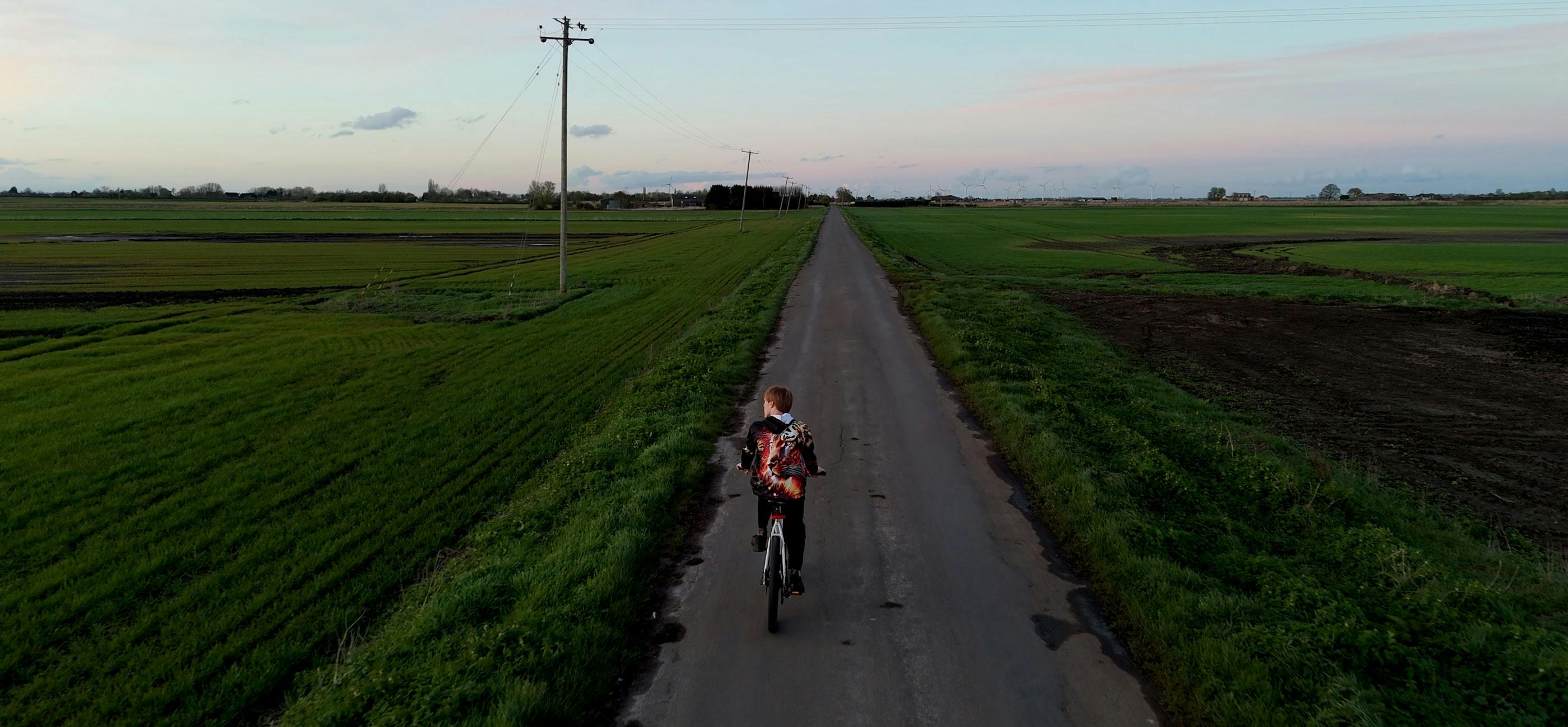 dan-canham.-brad-still.jpg A boy in a hoodie cycles down a long straight road between two flat fields in the Fens