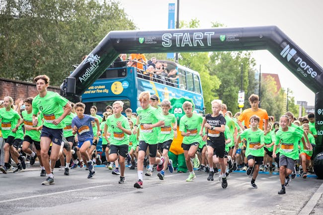 Group of children running from the start line while people on a parked bus cheer them on.